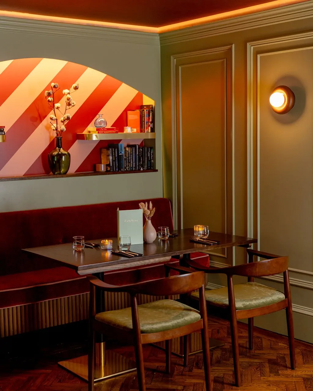 A cozy corner dining area in a restaurant featuring a deep red velvet banquette, dark wood tables, and olive green walls with decorative paneling, illuminated by warm ambient lighting.