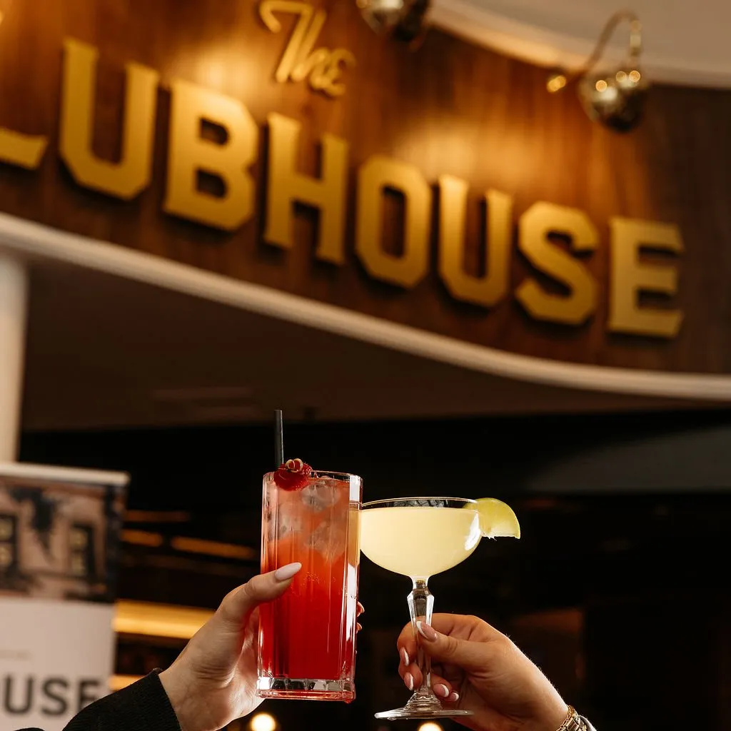 This close-up captures a celebratory moment, with two hands clinking vibrant cocktails against a softly blurred background. The backdrop features a prominent, dimensional "The CLUBHOUSE" sign in warm gold lettering on a rich, dark wood panel, illuminated by subtle sconces, suggesting an upscale yet inviting lounge or bar setting. The warm, ambient lighting creates a sophisticated and intimate atmosphere, perfect for evening gatherings.
