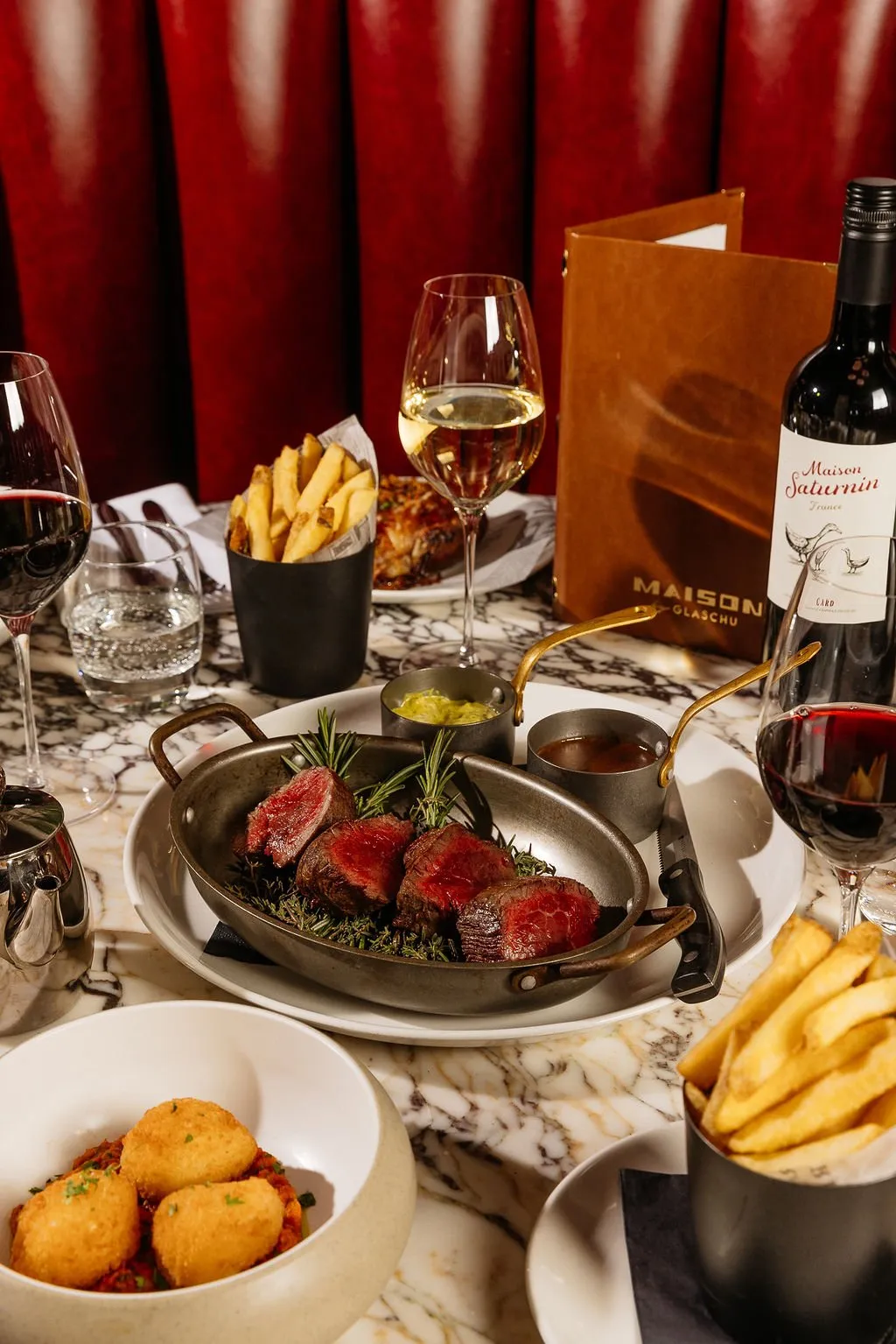 This close-up captures a luxurious dining experience, featuring a perfectly seared steak served in a rustic metal pan alongside golden fries and arancini, all presented on a veined marble tabletop. The rich, deep red upholstered banquette in the background, combined with soft, warm lighting, creates an intimate and sophisticated atmosphere, complemented by elegant wine glasses and a branded wine bottle.