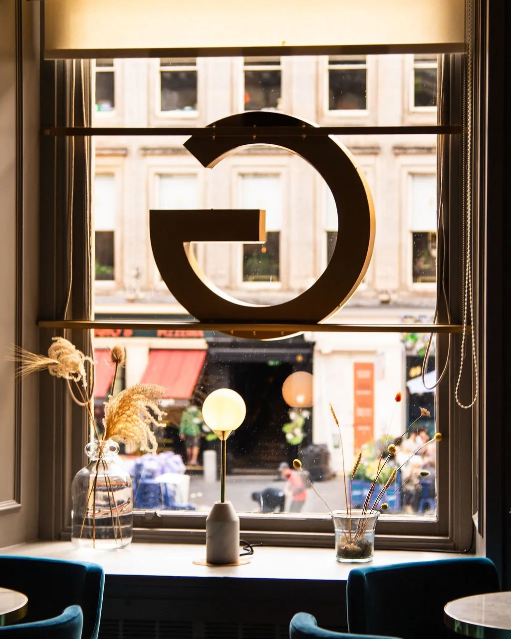 A cozy interior window scene looks out onto a bustling city street, featuring a prominent golden 'G' logo suspended within the window frame. The window sill is thoughtfully styled with a minimalist marble and brass globe lamp, flanked by clear glass vases holding dried grasses and seed pods, creating a warm and inviting vignette. Below, glimpses of plush teal velvet chairs and small round tables suggest a comfortable seating area, bathed in the soft glow of interior lighting contrasting with the muted daylight outside.