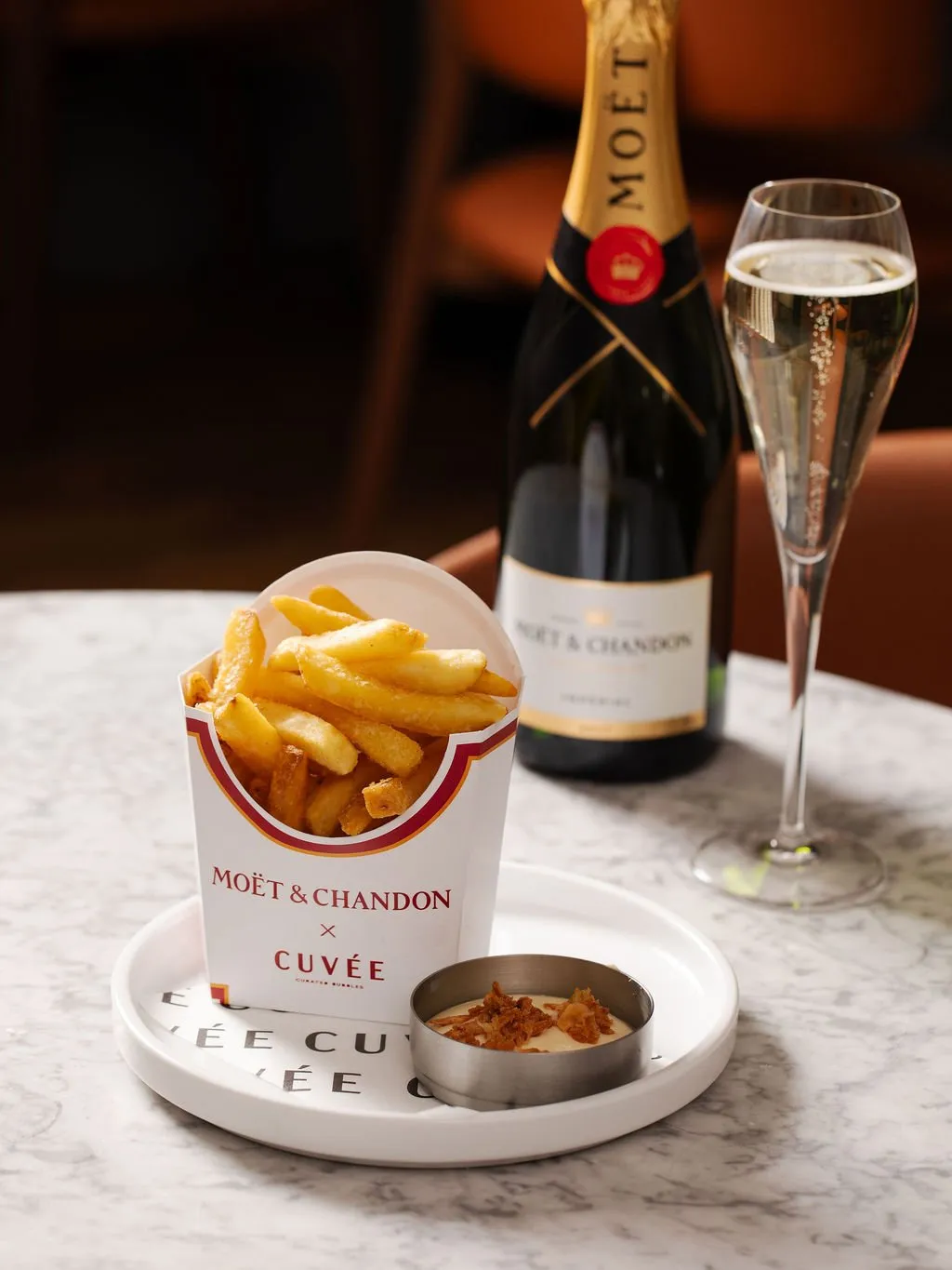 This close-up shot captures a luxurious yet casual pairing on a polished white marble tabletop. A branded Moët & Chandon x Cuvée paper bucket overflows with golden fries, accompanied by a small metal dish of creamy sauce topped with crispy elements. In the background, a chilled bottle of Moët & Chandon champagne stands beside an elegant flute filled with sparkling bubbly, hinting at a sophisticated, warmly lit hospitality setting with rich, dark orange furnishings.