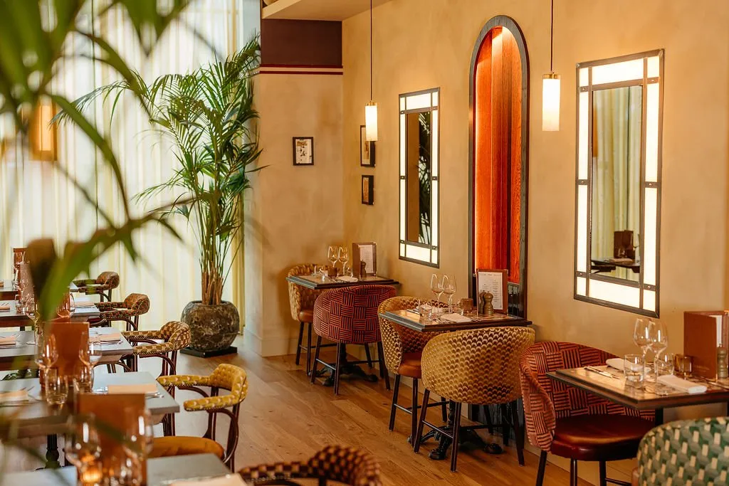 This inviting restaurant interior features a warm, sophisticated ambiance created by textured ochre walls and rich, patterned upholstery. Individual dining tables are arranged along a wall, each paired with unique chairs in mustard, burnt orange, and deep green geometric fabrics, complemented by dark metal bases. Ambient lighting from cylindrical pendant lights and striking backlit mirrors illuminates the space, highlighting a dramatic arched niche with warm wooden paneling, while a large potted palm adds a touch of natural elegance.