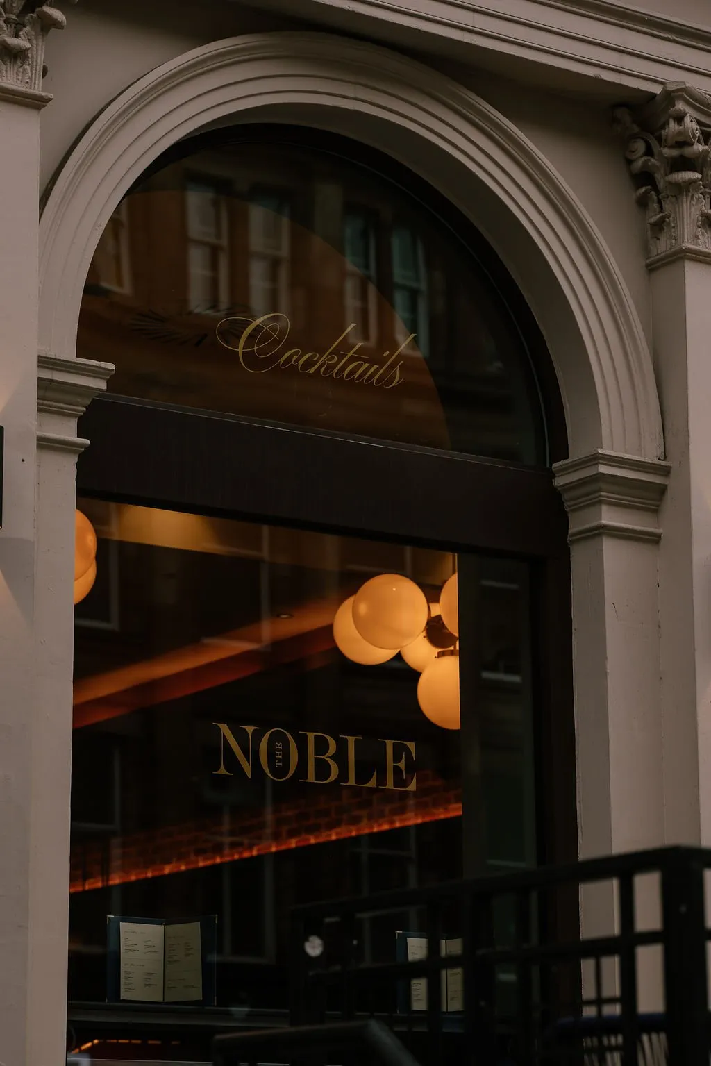 A close-up shot of an elegant arched entrance to 'The Noble' bar, featuring golden signage and warm spherical lighting visible through the glass.