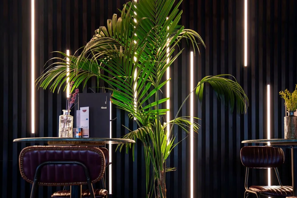 The scene captures an intimate corner of a modern hospitality space, featuring a striking dark slatted wall punctuated by vertical LED light strips that create a warm, ambient glow. A lush palm plant adds a vibrant natural element, partially obscuring the lighting and a small round table set with menus and a ribbed glass vase. The foreground reveals a comfortable, dark burgundy tufted leather chair with a metal frame, suggesting a sophisticated and inviting atmosphere.