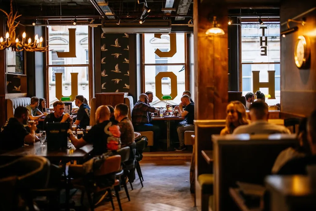This inviting hospitality space features a warm, rustic-industrial aesthetic, with dark wood paneling, exposed ceilings, and a mix of banquette and table seating. Ambient lighting from an antler chandelier, track lights, and wall sconces creates a cozy, social atmosphere, complemented by natural light filtering through large windows adorned with bold yellow typography. The layout encourages lively interaction, with patrons enjoying drinks and conversation across various comfortable zones.