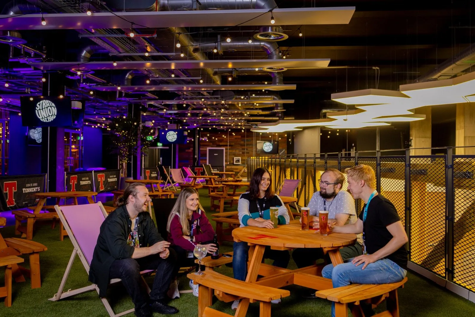 This vibrant, industrial-chic indoor space transforms a large hall into a playful, park-like social hub. Exposed ductwork and concrete ceilings are softened by string lights and geometric LED panels, casting a dynamic purple glow over artificial turf flooring. The layout features numerous rustic wooden picnic tables and colorful fabric deck chairs, fostering a relaxed, communal atmosphere perfect for casual gatherings and lively interaction.