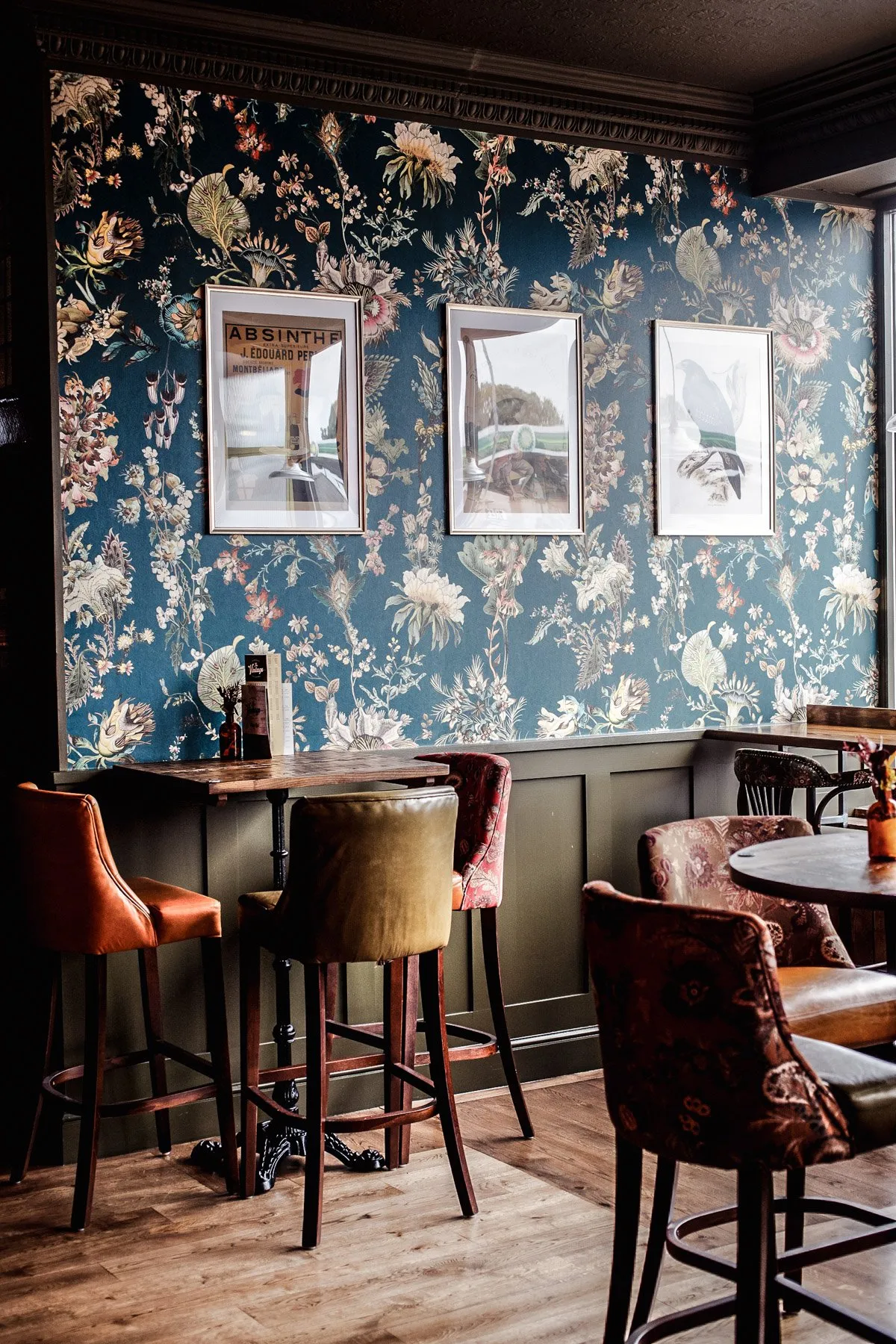 This inviting interior showcases a cozy corner of a hospitality space, likely a pub or bar, defined by a dramatic dark teal floral wallpaper with intricate botanical patterns. Below, dark olive-green wainscoting grounds the space, topped by a richly detailed dark wood ceiling trim. The layout features high-top wooden tables with a mix of seating, including warm orange leather bar stools, olive-green upholstered stools, and patterned fabric chairs, all contributing to a warm, intimate, and eclectic atmosphere under ambient lighting.