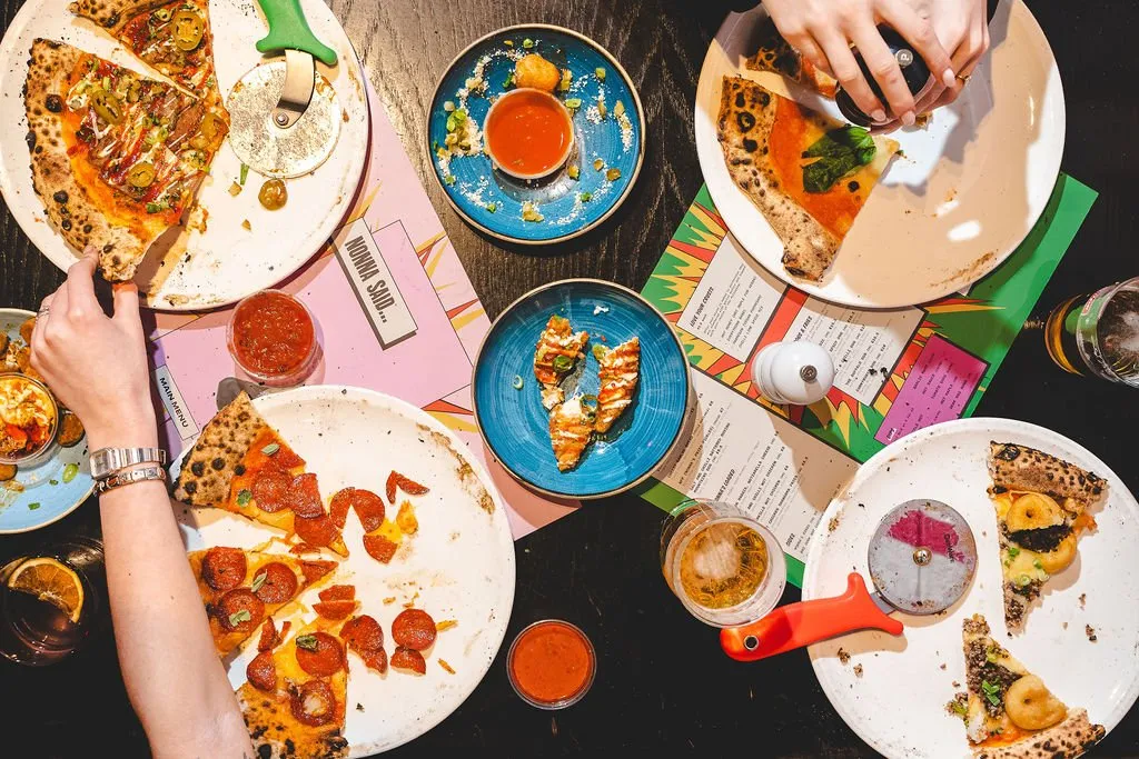 A vibrant overhead shot captures a lively shared meal at a contemporary pizzeria, showcasing multiple partially eaten pizzas with diverse toppings, alongside colourful side dishes and drinks. The dark tabletop is accented by playful, graphic paper menus in bold pinks, greens, and yellows, creating a casual and energetic atmosphere. Hands are actively engaged in cutting and serving pizza, highlighting the communal and interactive dining experience.