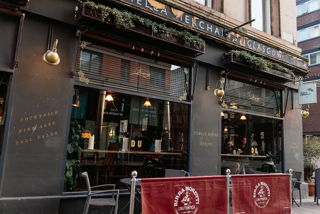 This exterior shot captures the inviting facade of a classic public house, "The Duke's Umbrella," featuring a dark charcoal-grey exterior with elegant gold script lettering and ornate brass sconces. Large windows reveal a warm, dimly lit interior with wooden furniture, creating a cozy and traditional atmosphere. Lush greenery in planter boxes above the windows adds a touch of natural charm, while red branded barriers hint at outdoor seating.