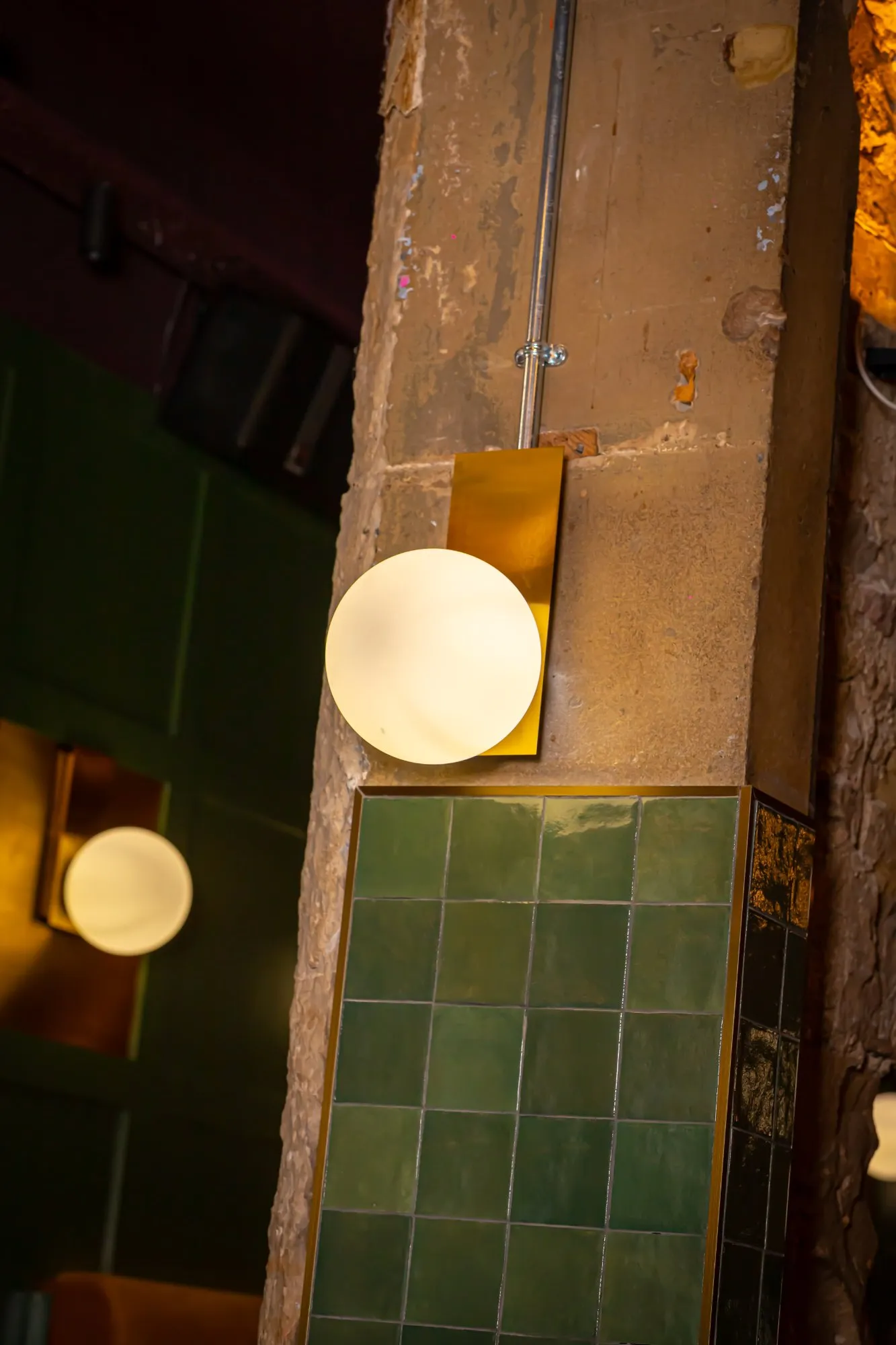 This close-up reveals a richly textured interior corner, featuring a distressed concrete pillar adorned with a modern brass and opal glass spherical wall sconce. Below the sconce, a section of vibrant, square green glazed tiles with dark grout adds a pop of colour and a touch of classic elegance, framed by a brass trim. The warm, ambient glow from the fixture creates an intimate and sophisticated mood, highlighting the interplay between raw industrial elements and refined design details. Hints of a dark green wall and possibly a curved banquette are visible in the soft background, suggesting a hospitality setting.