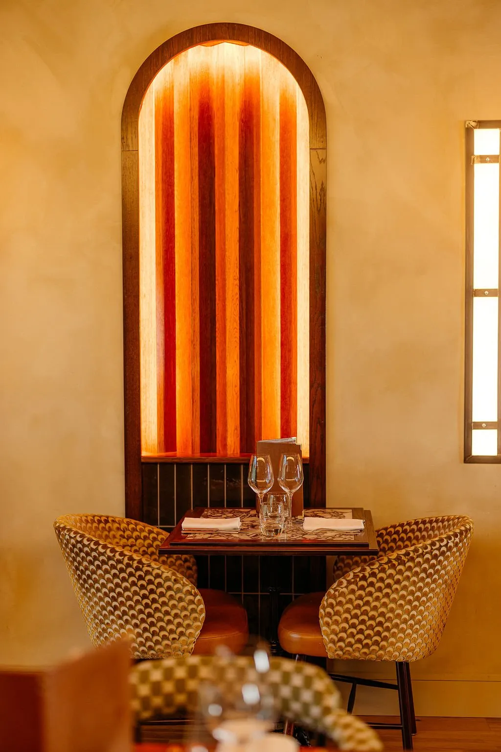 This intimate dining nook features a small square table set for two, flanked by two striking armchairs upholstered in a gold and cream geometric pattern. The focal point is a backlit arched niche with vertical wooden slats displaying a warm orange-to-red gradient, complemented by a subtle rectangular wall sconce. The overall mood is warm, sophisticated, and inviting, with a rich material palette of wood, patterned fabric, and plaster.