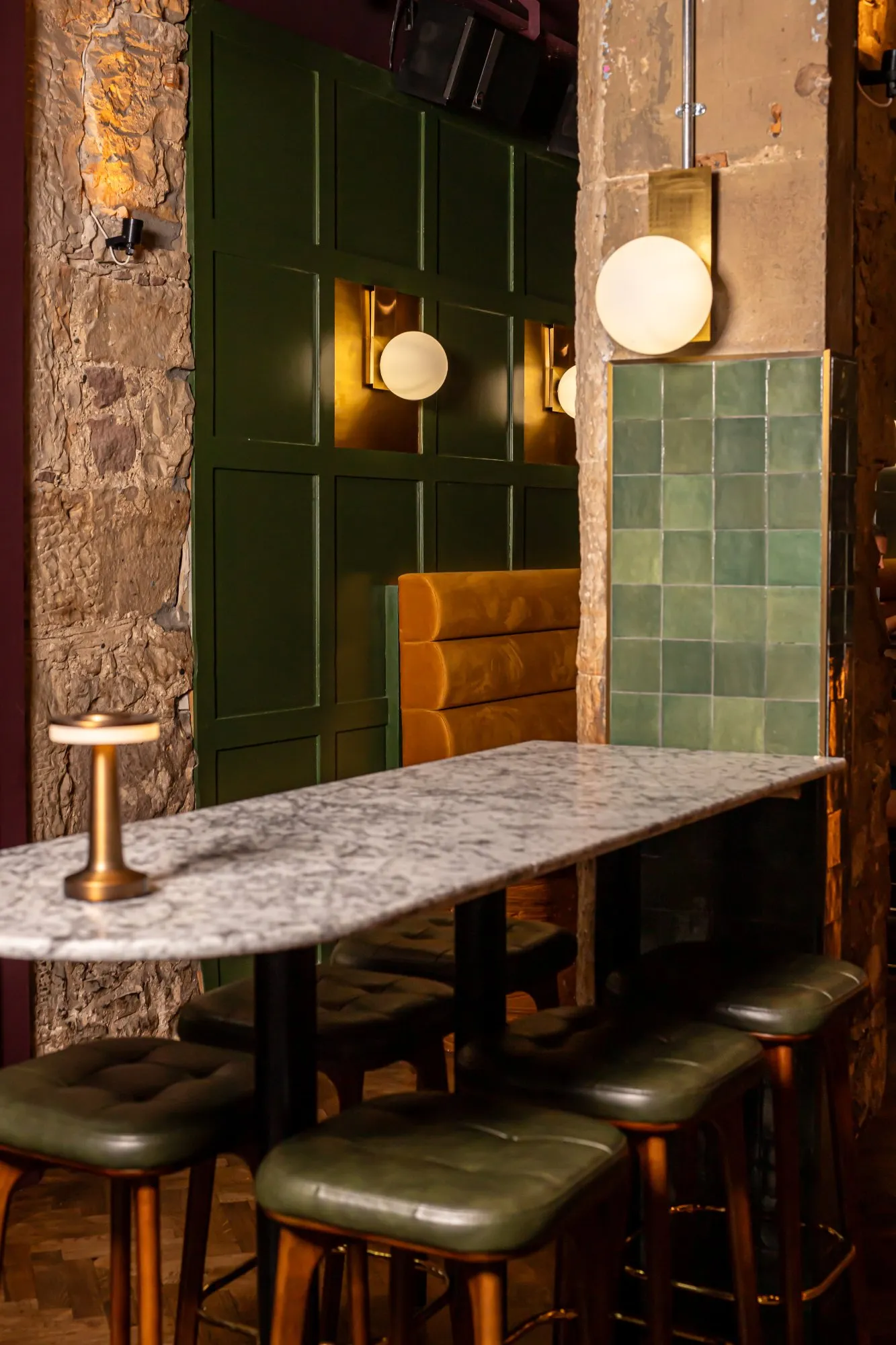 This intimate hospitality space features a rich interplay of textures and warm lighting, creating a sophisticated and inviting atmosphere. A high-top marble table with dark green leather stools provides casual seating, complemented by a plush mustard-yellow banquette set against a dark green paneled wall. Exposed rough stone and distressed columns contrast with elegant brass spherical sconces and subtle green square tiles, all bathed in a moody, inviting glow.