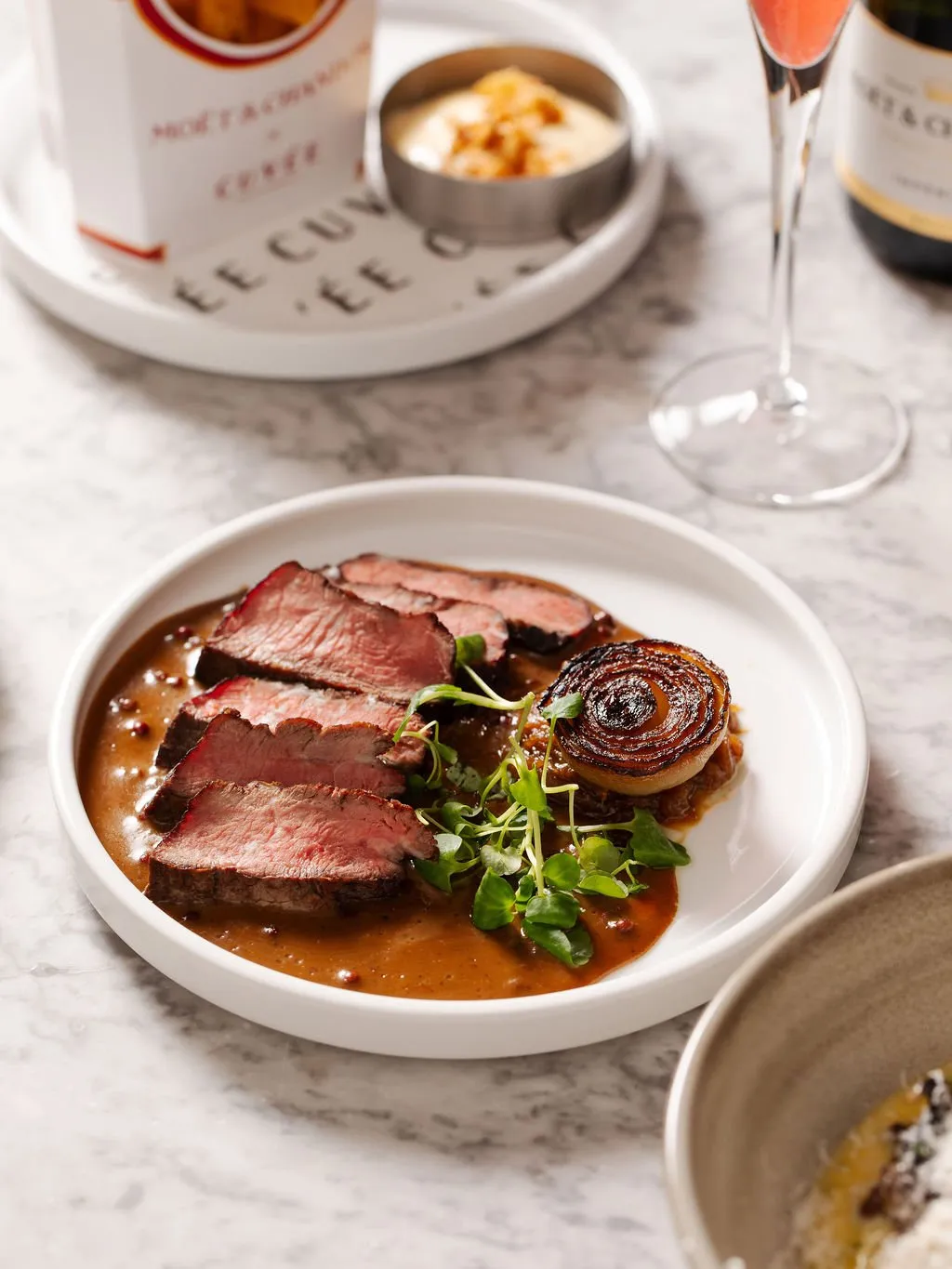 A close-up shot captures a meticulously plated main course featuring perfectly seared, medium-rare sliced steak with a rich brown sauce, garnished with a caramelized roasted onion half and fresh watercress, all presented on a minimalist white ceramic plate. The dish rests on a luxurious white marble tabletop, accompanied by a delicate champagne flute, a bottle of champagne, and a side of crispy fries with dipping sauce in branded packaging, suggesting an upscale and celebratory dining experience. The bright, natural lighting highlights the textures and vibrant colours of the gourmet food and elegant setting.
