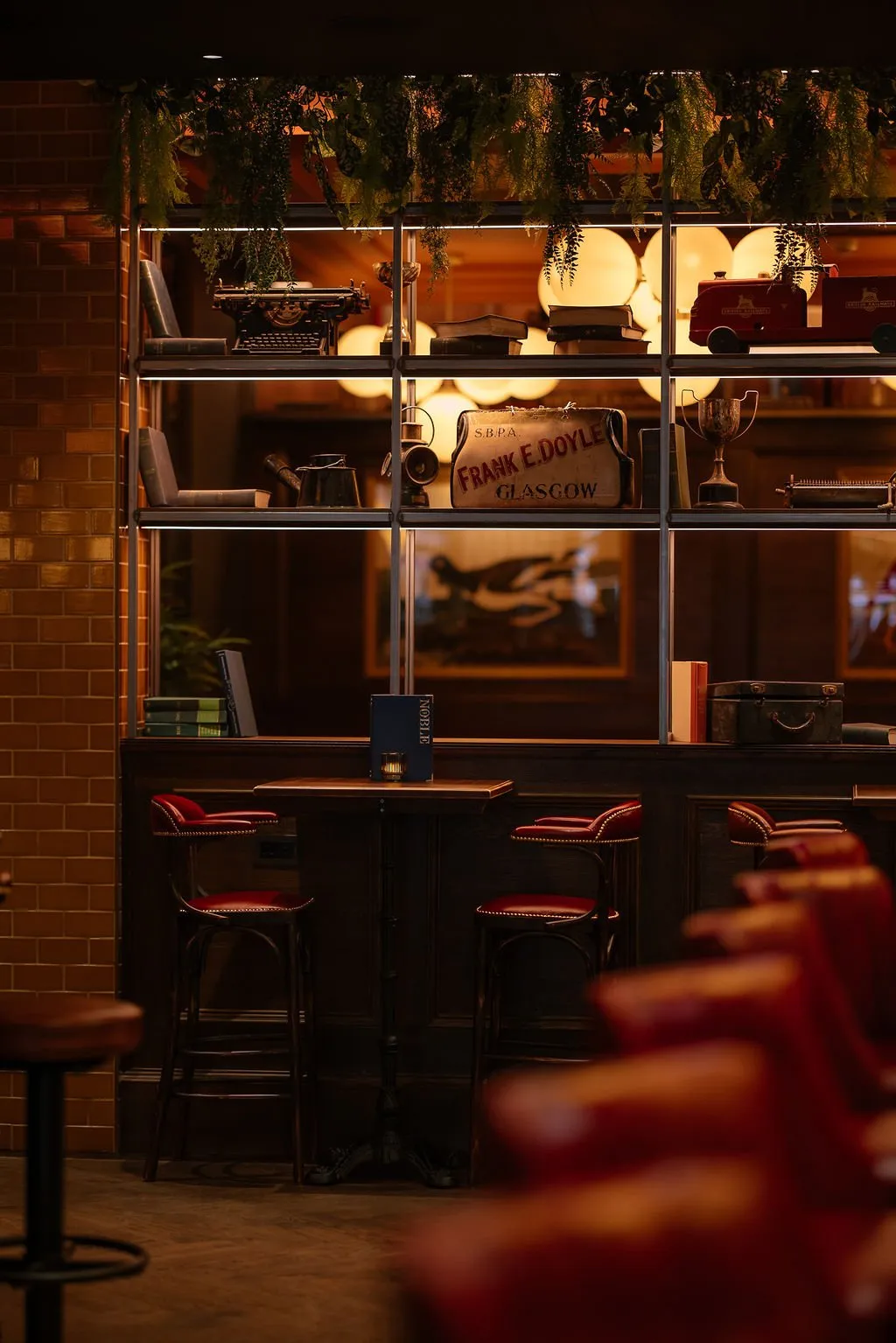 A warmly lit interior shot of a bar or lounge area featuring a display shelf filled with vintage curiosities above a dark wood bar and red leather stools.