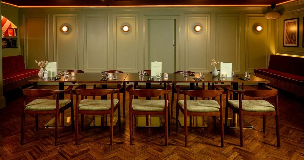 A sophisticated dining room features sage green paneling, a long wooden table set for a meal, and deep red banquette seating under warm ambient lighting.