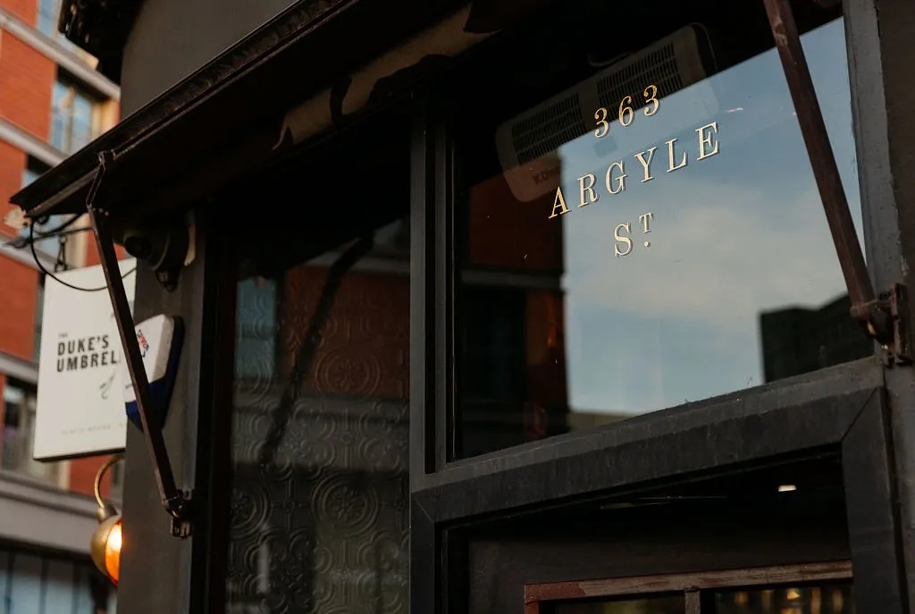 This close-up captures the inviting exterior of a hospitality venue, featuring a dark, weathered facade with prominent gold-leafed address numbers and street name, '363 ARGYLE ST', applied directly to the large glass window. A custom white hanging sign, 'THE DUKE'S UMBREL', and a warm, glowing brass wall sconce add character, while reflections in the glass hint at both the evening sky and a subtly patterned interior wall. The overall effect creates a moody, sophisticated, and welcoming urban entrance at dusk.