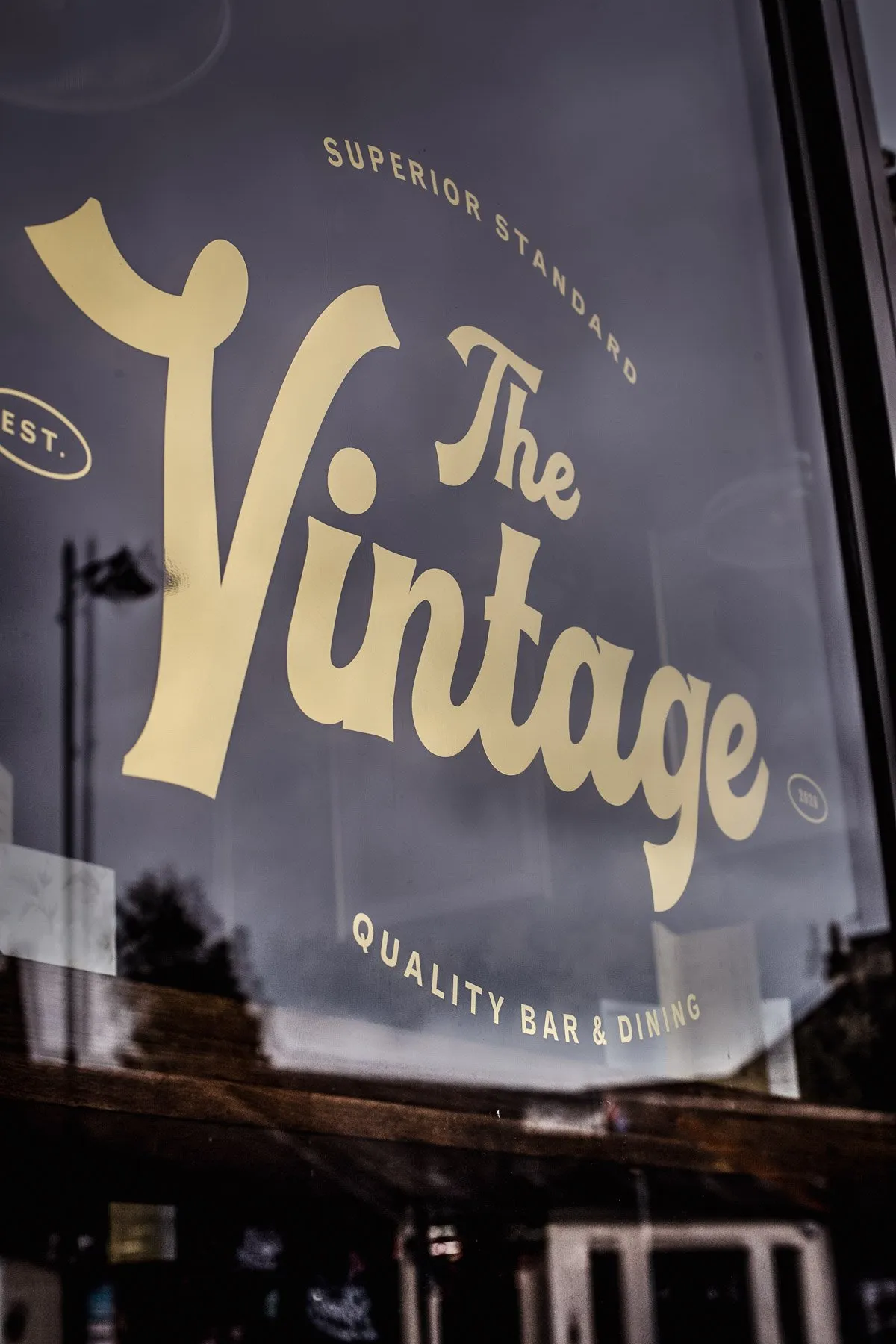This image captures a close-up of a hospitality venue's shopfront window, showcasing its elegant brand identity. Gold vinyl lettering, featuring a sophisticated script for "The Vintage" and classic sans-serif text for "Superior Standard" and "Quality Bar & Dining," is prominently displayed against the dark, reflective glass. The window subtly mirrors the blurred outdoor environment, adding depth and context to the inviting, established aesthetic.