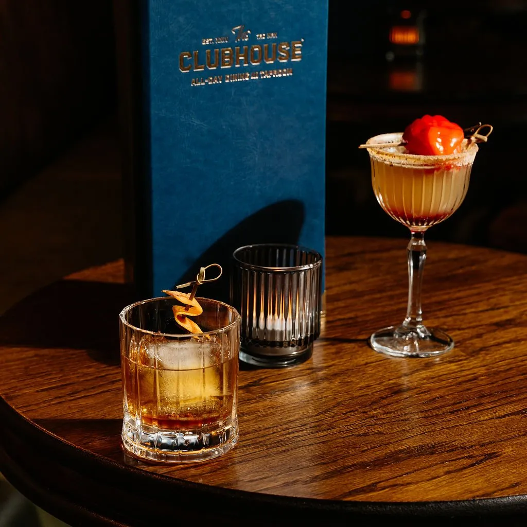 This close-up shot captures an intimate, sophisticated evening setting, highlighting two artfully crafted cocktails on a rich, dark stained wooden table. A deep blue menu with elegant gold foil lettering stands prominently in the background, casting a soft shadow. The warm, directional lighting creates a moody ambiance, emphasizing the intricate details of the fluted glassware and the vibrant garnishes, suggesting a premium hospitality experience.