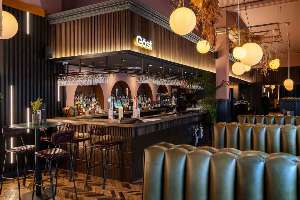 This sophisticated bar and dining space features a long, dark fluted bar counter with a lighter wood top, backed by arched niches displaying an extensive liquor selection against a soft pink wall. Opposite the bar, rows of plush olive-green tufted leather banquettes offer comfortable seating, complemented by dark metal and brown leather bar stools and high-top tables with dark stone tops. The warm, inviting atmosphere is created by numerous large spherical opal pendant lights and vertical linear LEDs, highlighting the rich textures of dark slatted walls, herringbone wood flooring, and ornate ceiling cornices.
