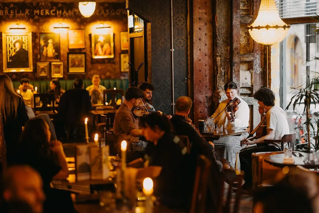 This interior shot captures a vibrant, dimly lit pub or bar, exuding a cozy and traditional atmosphere. Dark, richly textured wallpaper adorned with a gallery wall of framed portraits and illuminated by picture lights sets a classic backdrop, complemented by distressed wooden columns and a deep green banquette. Warm, ambient lighting from ornate chandeliers, wall sconces, and numerous flickering candles on dark wooden tables creates an intimate mood, where patrons enjoy live folk music from a group of musicians.