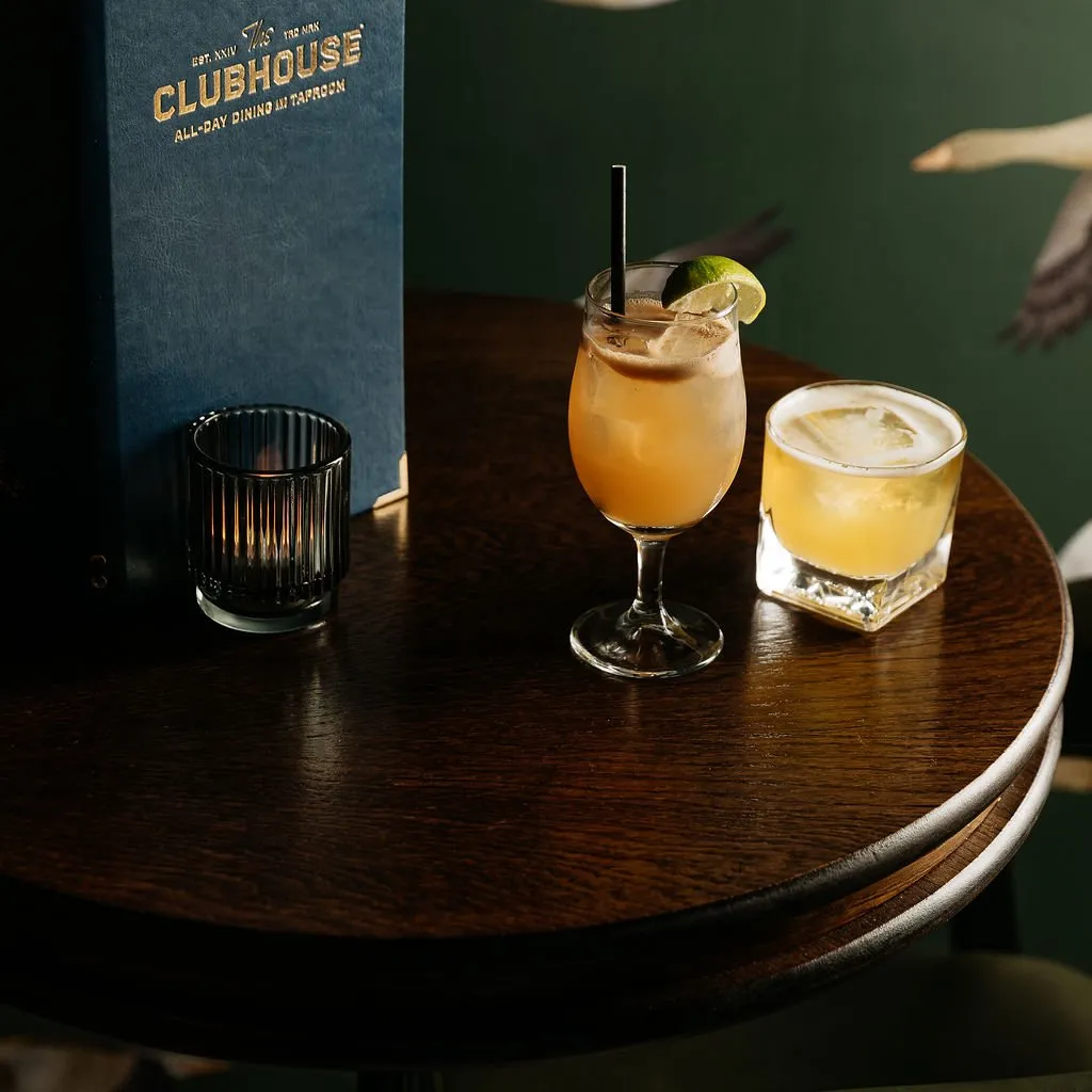 This intimate close-up captures a dark, richly stained round wooden table set for evening drinks. Two inviting cocktails, a ribbed glass candle holder casting a warm glow, and a deep blue menu with gold foil lettering are prominently displayed. The background reveals a sophisticated dark green wallpaper featuring a subtle, elegant bird motif, contributing to a cozy and refined atmosphere.