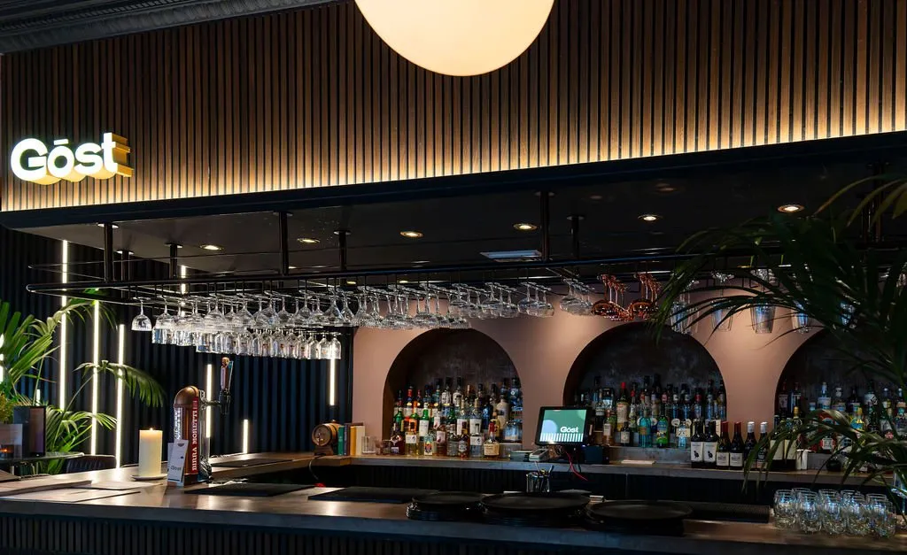 This sophisticated bar features a striking design with a dark, slatted bar front and a contrasting light wood slat wall paneling above, dramatically backlit to highlight its texture. Overhead, a large spherical pendant light casts a soft glow, complementing the warm ambient lighting from integrated vertical LED strips and recessed spotlights. The back bar showcases a series of elegant arched recesses in a muted pink hue, displaying an array of bottles beneath a sleek overhead glass rack, all contributing to a modern yet inviting atmosphere.