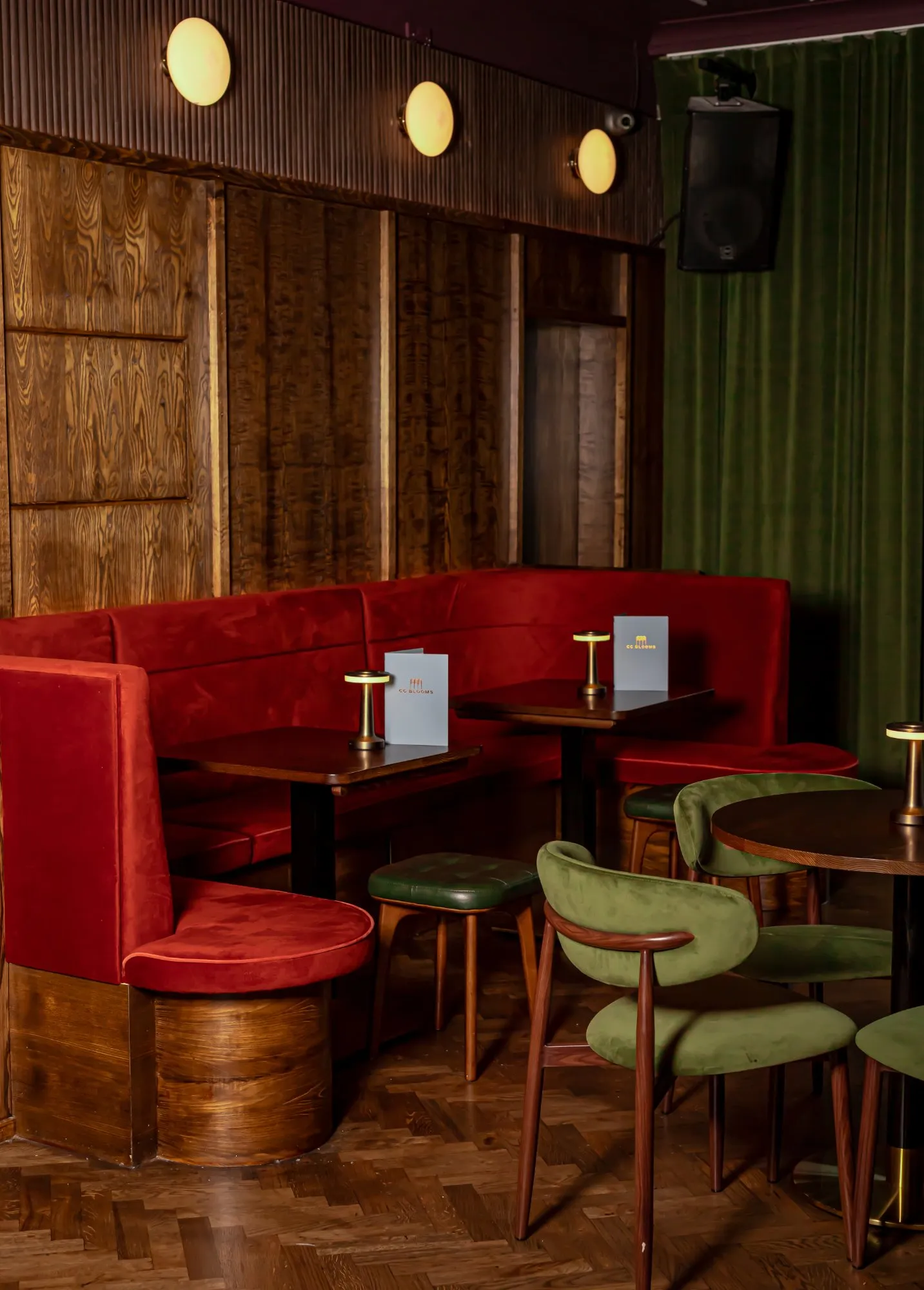 This intimate bar or lounge area features a rich material palette, creating a warm and sophisticated atmosphere. A long, curved banquette upholstered in deep red velvet anchors the space against dark, richly grained wood paneling, complemented by olive green velvet chairs and small leather stools. Warm, spherical wall sconces and modern brass table lamps cast a soft glow, highlighting the herringbone wood floor and deep green velvet curtains, evoking a luxurious, retro-inspired ambiance perfect for evening gatherings.