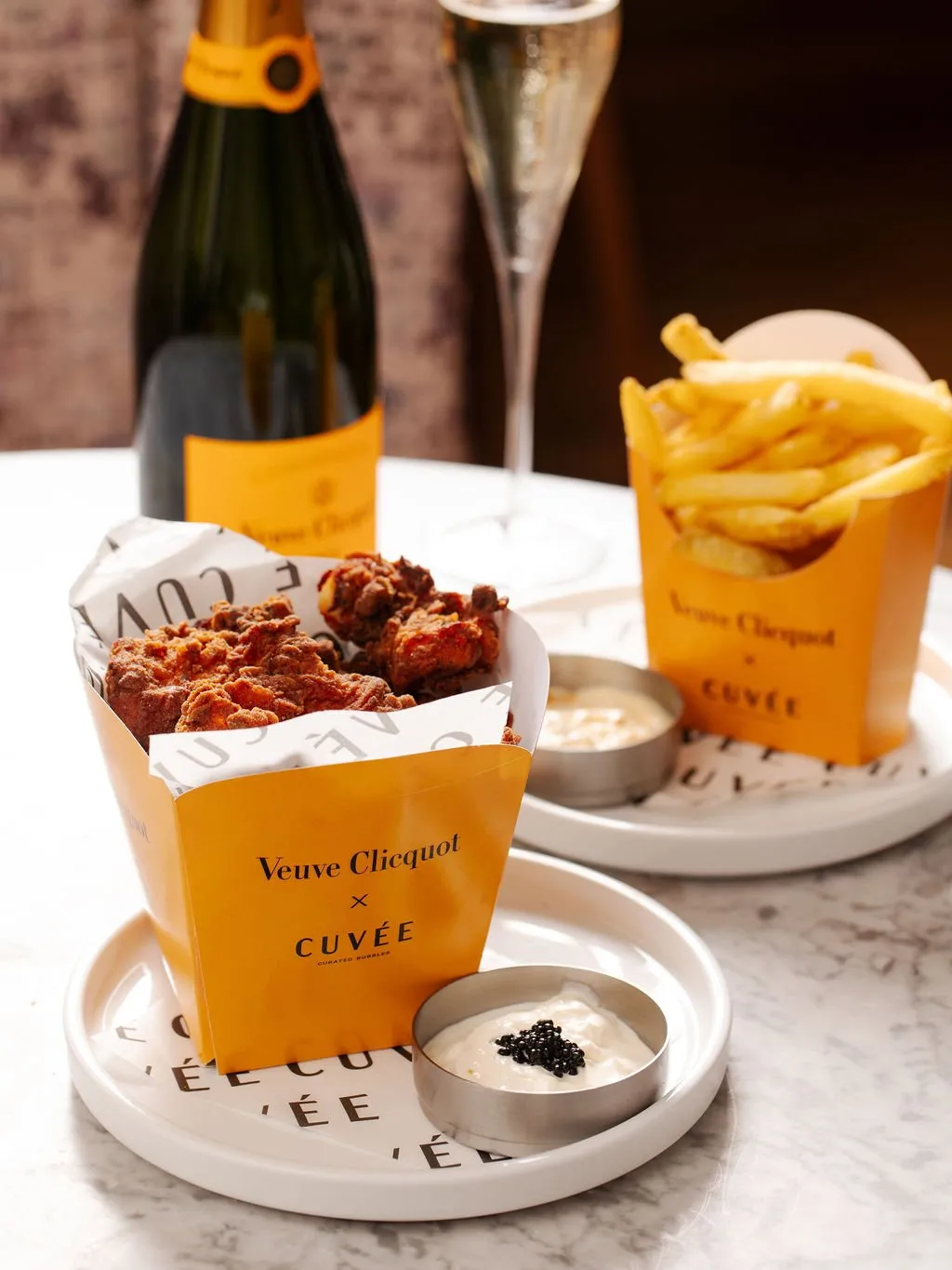 This close-up shot captures a luxurious yet casual dining experience, featuring gourmet fried chicken and crispy fries served in distinctive branded Veuve Clicquot x Cuvée packaging. A bottle of champagne and a flute stand in the softly blurred background, suggesting a celebratory mood. The food is elegantly presented on white ceramic plates atop a subtly veined marble table, with a notable detail of black caviar garnishing one of the dipping sauces, elevating the indulgence.