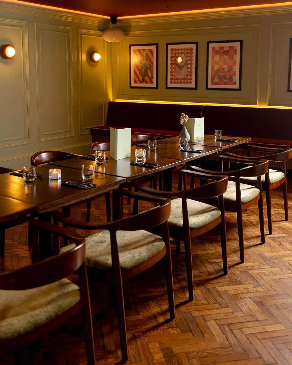 A warm and inviting dining area features a long wooden table set for guests, complemented by a deep burgundy banquette and sage green paneled walls.