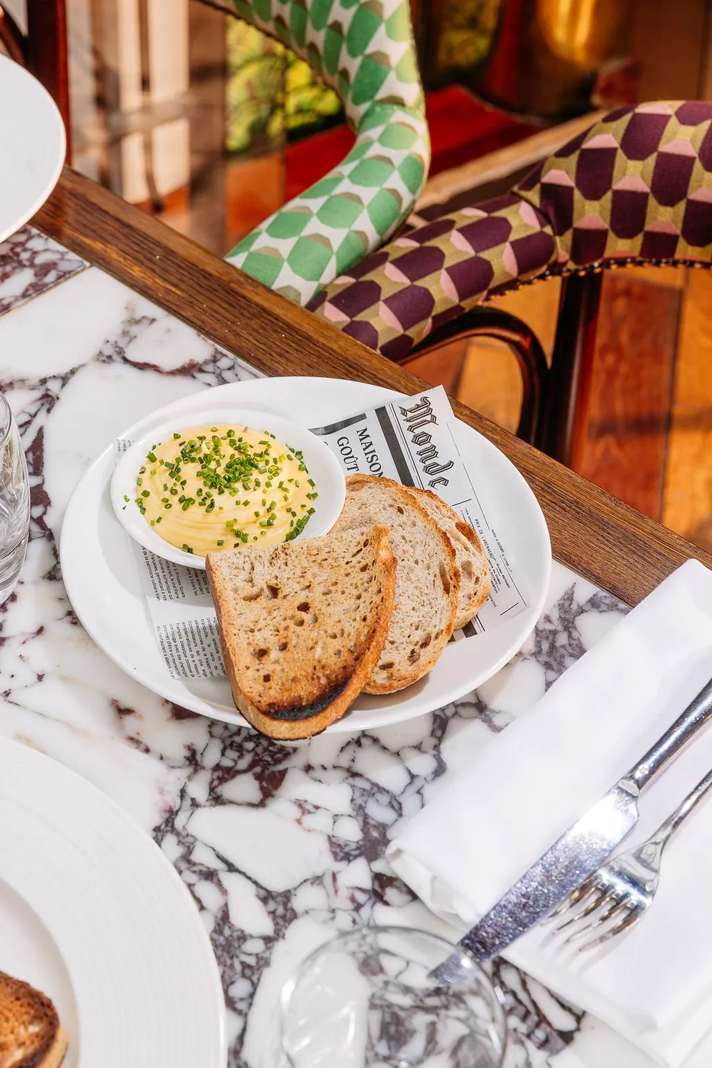 This close-up shot captures a sophisticated dining table setting, featuring a striking white marble tabletop with rich burgundy veining. In the background, a classic wooden chair is upholstered with two distinct geometric patterns in vibrant green and plum hues, adding a playful yet elegant touch. Bright natural light illuminates the scene, highlighting a plate of toasted bread and a bowl of chive-topped spread, creating an inviting and chic atmosphere.