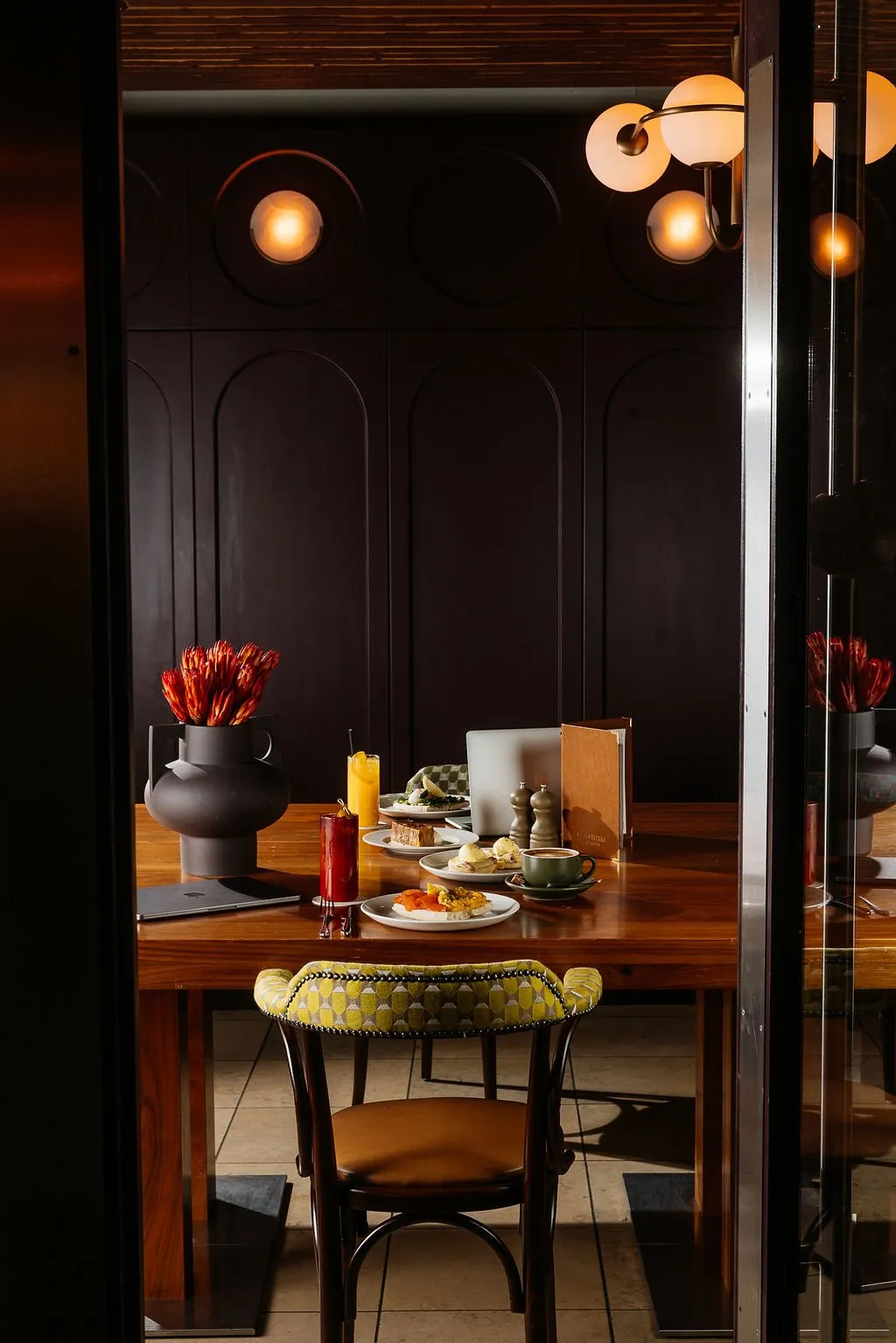 The image captures an intimate dining setting within a sophisticated hospitality space, framed by a dark doorway. A rich wooden table is laden with a vibrant breakfast spread, flanked by a striking bentwood-style chair featuring a geometric chartreuse upholstery and brass stud detailing. The backdrop is a deep plum-toned paneled wall with elegant arched lower sections and integrated circular wall sconces, complemented by a multi-globe pendant light, all contributing to a warm, inviting, and subtly luxurious atmosphere.