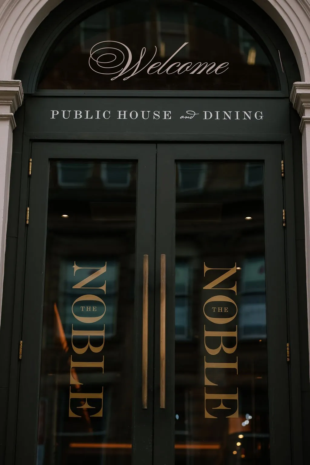 A close-up exterior shot of a dark green double door entrance to "The Noble Public House and Dining," featuring elegant gold lettering and brass hardware.