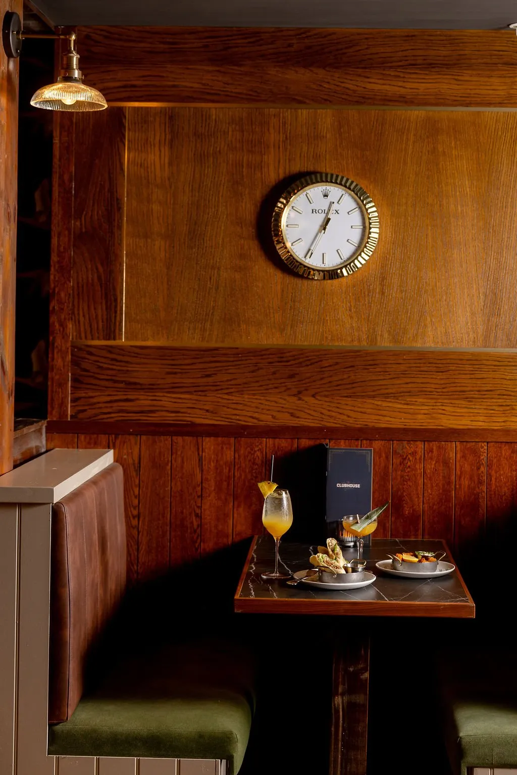 This intimate booth setting features rich, dark wood paneling with subtle brass accents, creating a classic and sophisticated atmosphere. A plush banquette with a brown leather back and olive green velvet seat is paired with a dark marble-topped table, laden with cocktails and small plates, all bathed in the warm glow of a fluted brass wall sconce. The iconic Rolex wall clock adds a touch of timeless luxury to the clubby ambiance.