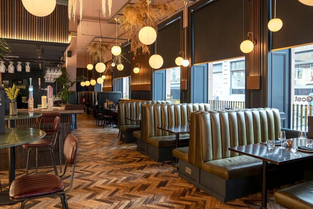 This sophisticated hospitality interior showcases a long row of olive-green channel-tufted booth seating set against dark, vertically paneled walls, complemented by a rich herringbone wood floor. A constellation of brass and opal globe pendant lights, interspersed with dramatic dried floral ceiling installations, casts a warm, inviting glow throughout the space. The layout includes a well-appointed bar area with high-top seating, while large windows with dark roller blinds offer an urban backdrop. The overall mood is intimate and elegant, blending contemporary design with classic comfort.