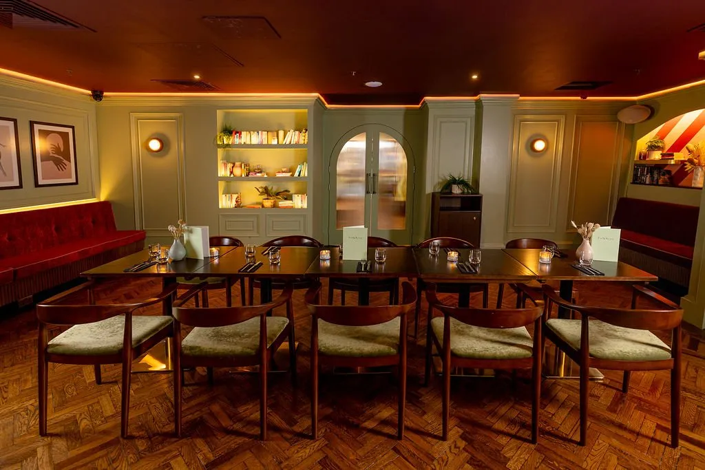 A stylish and inviting restaurant dining area features a long communal table, plush red banquette seating, and illuminated decorative shelving.