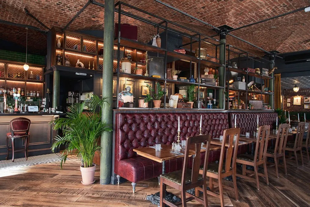 This inviting hospitality space features a rich, eclectic design with a vaulted exposed brick ceiling and distressed dark wood flooring. A long, tufted deep red leather banquette runs alongside a series of dark wooden tables and chairs, facing a well-stocked bar with dark wood paneling and green subway tiles. Overhead, an elaborate dark metal shelving system, adorned with an array of vintage curios and illuminated by warm strip lighting, creates a library-like atmosphere, complemented by ambient pendant lights and natural light near the bar. Dark green structural columns and a large potted plant add to the layered, sophisticated mood.