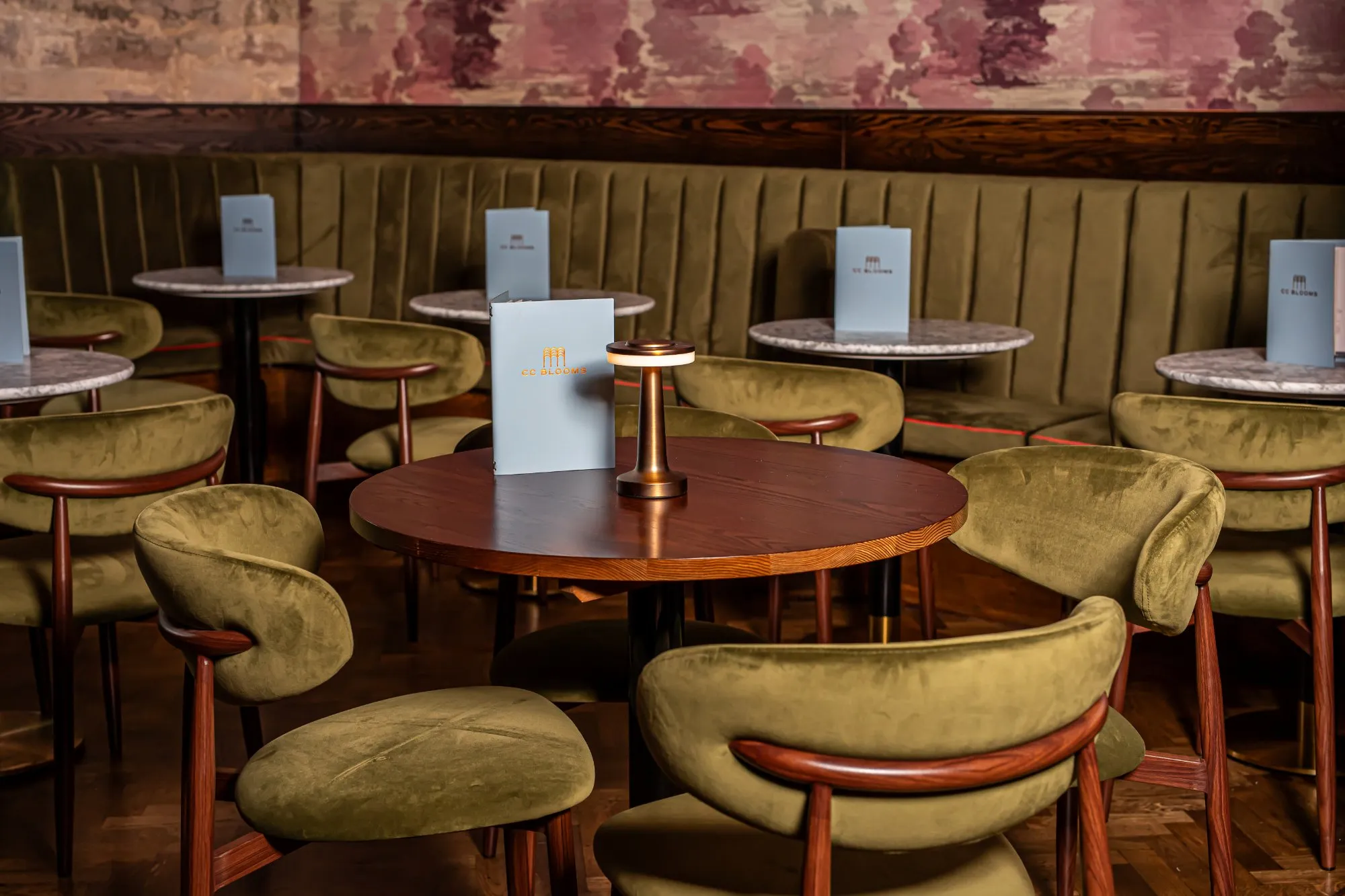 A sophisticated and intimate dining or lounge area features a long, channel-tufted olive green velvet banquette running along a wall adorned with a faded, abstract floral wallpaper in muted pinks and browns. Dark wood round tables, some with marble tops, are paired with elegant olive green velvet chairs featuring distinctive curved dark wood frames. The space is softly illuminated by modern, portable bronze table lamps, creating a warm and inviting evening atmosphere perfect for a high-end hospitality setting.