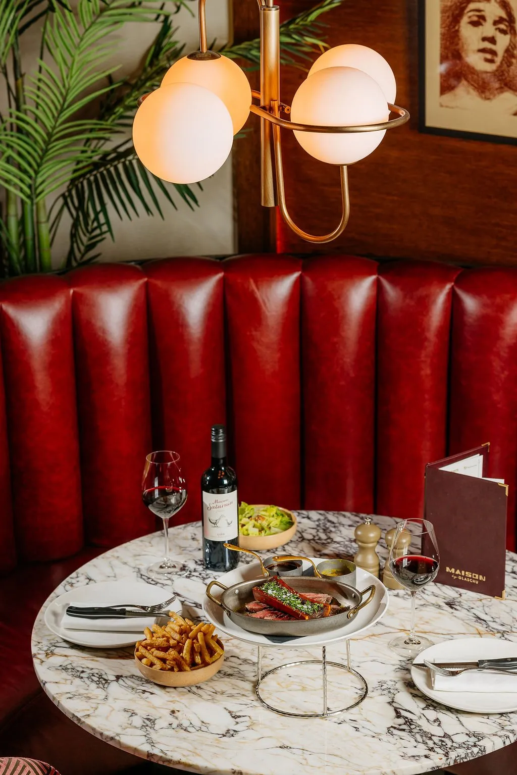 This intimate restaurant setting features a round white marble-top table set for two, backed by a luxurious deep red channel-tufted banquette. Warm, ambient light emanates from a modern brass pendant fixture with spherical opal glass shades, creating a cozy and inviting atmosphere. The table is elegantly appointed with a steak dish, a bottle of red wine, and accompanying sides, suggesting a refined yet comfortable dining experience.