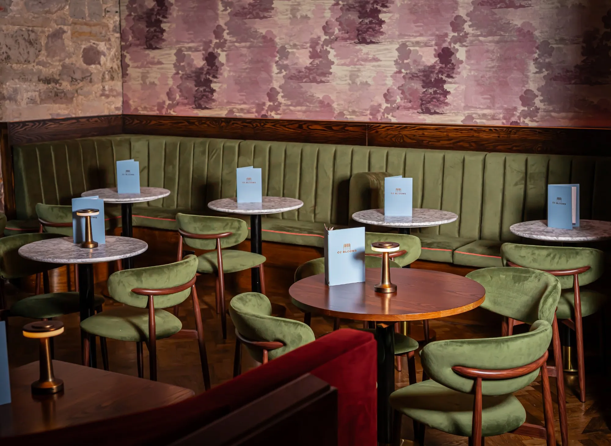 This intimate hospitality space features a rich material palette and moody lighting, creating a sophisticated and inviting atmosphere. A long, curved olive green velvet banquette with subtle coral piping lines one wall, complemented by individual olive green velvet chairs with dark wood frames. Round tables with both white marble and dark wood tops are illuminated by modern brass-toned portable lamps, set against a dramatic pink and purple abstract wallpaper and an exposed stone wall.