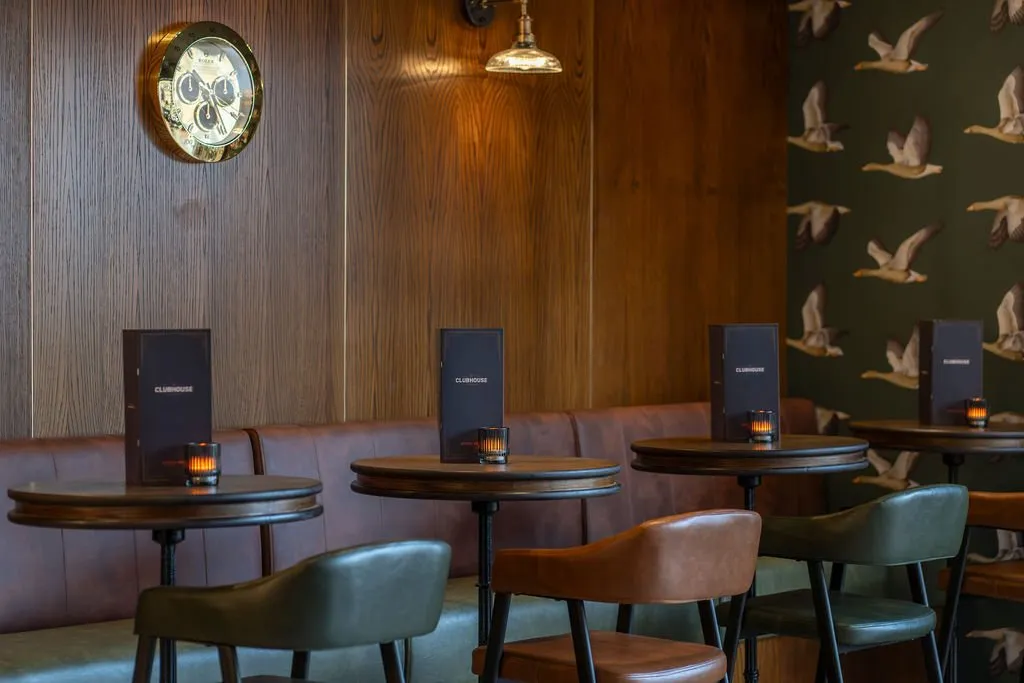 This intimate lounge area features a rich material palette, combining dark wood paneling with a deep green wallpaper adorned with a flying geese motif. A long, tufted brown leather banquette provides continuous seating, complemented by individual round tables and mixed leather chairs in shades of brown and olive. Warm, inviting light emanates from a brass wall sconce and small votive candles, creating a sophisticated and cozy atmosphere perfect for evening gatherings.