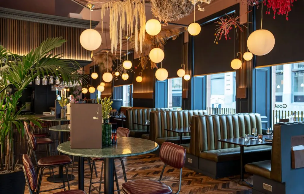 This sophisticated restaurant interior features a rich material palette and warm, ambient lighting. Deep olive-green ribbed banquettes line the windows, complemented by dark reddish-brown leather chairs and green marble-topped tables, all set against a striking herringbone wood floor. The ceiling is adorned with numerous spherical pendant lights at varying heights and dramatic dried botanical installations, while fluted dark wood paneling adds texture to the walls, creating an intimate and inviting atmosphere.