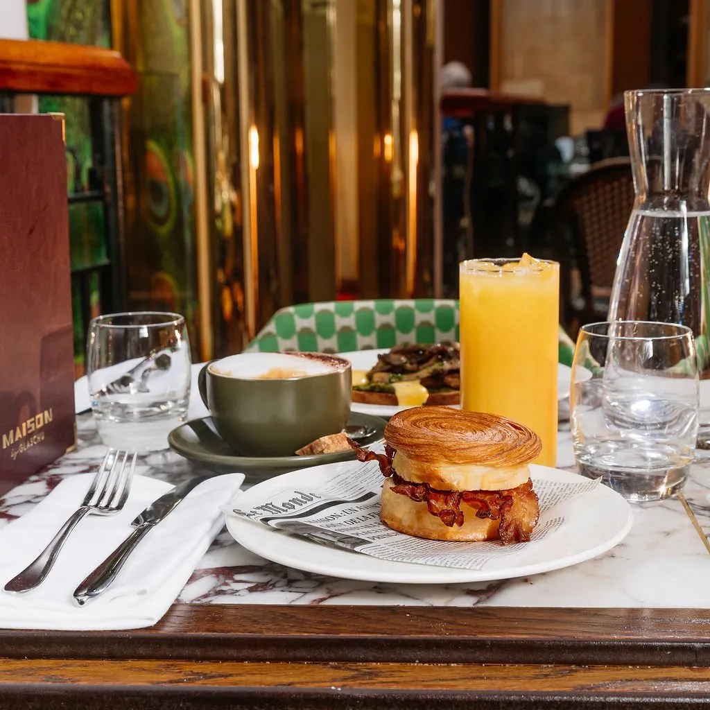 This close-up shot captures a vibrant breakfast or brunch setting on a polished white marble table with dark wood trim. The scene is bright and inviting, featuring a croissant sandwich, a cappuccino in a deep green ceramic cup, orange juice, and water. In the background, highly reflective brass or gold panels create a luxurious backdrop, complemented by a distinctive green geometric patterned chair and a classic woven bistro chair, hinting at a sophisticated yet comfortable cafe or restaurant interior.