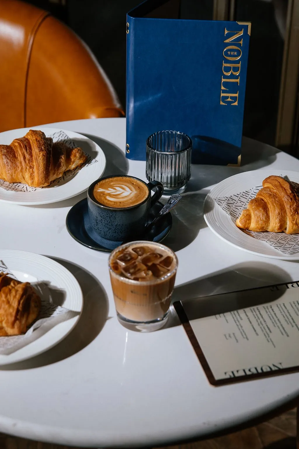 A close-up shot of a white table set with coffee, iced coffee, and croissants, featuring a blue branded menu and a glimpse of a warm-toned leather chair in the background.