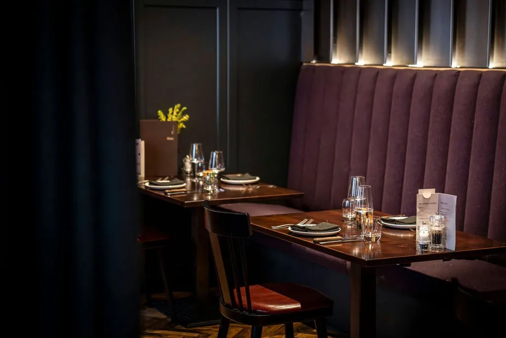 This intimate dining area features a sophisticated palette of deep aubergine and dark wood, creating a warm and inviting atmosphere. A long, channel-tufted banquette in rich velvet runs along a dark paneled wall, complemented by individual dark wood chairs with reddish-brown leather seats. Subtle uplighting above the banquette casts a soft glow, highlighting the texture and enhancing the cozy, upscale mood for an evening meal. The tables are meticulously set with glassware, dark napkins, and menus, ready for service.