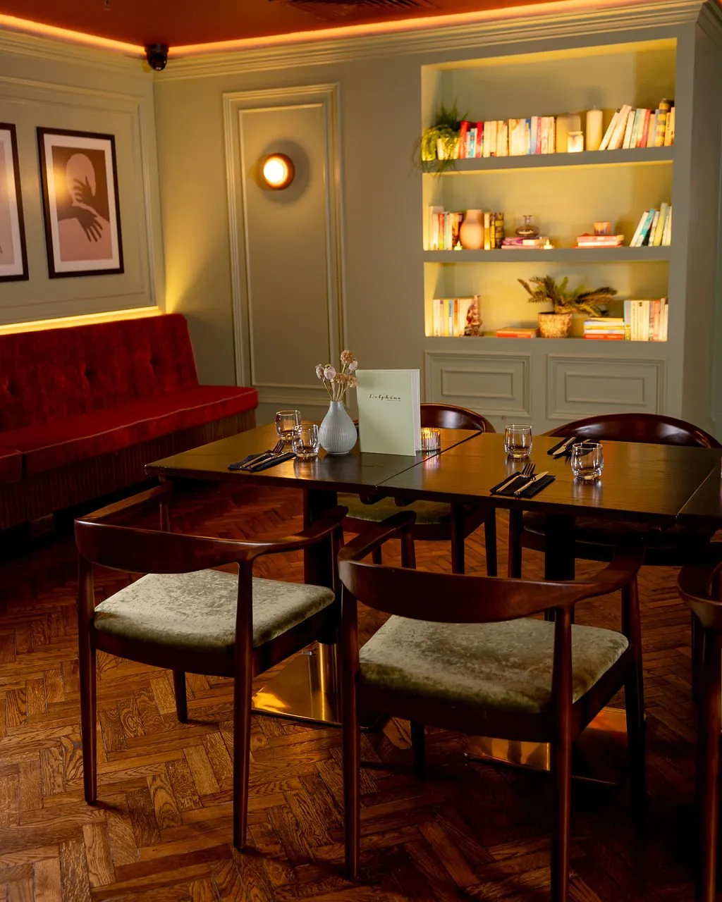 This inviting dining space features a rich palette of sage green walls, a deep red velvet banquette, and warm wooden furniture, illuminated by ambient and accent lighting.