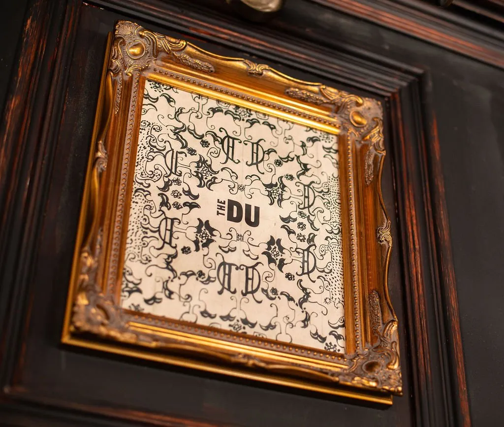This close-up shot features an ornate, gilded baroque-style frame holding a sign with a distinctive black-and-white pattern. The pattern incorporates stylized letters and organic flourishes, with "THE DU" prominently displayed in a modern sans-serif font. The frame is mounted on a dark, richly paneled wooden wall, suggesting a classic, perhaps opulent, interior with a blend of traditional and contemporary elements. The lighting highlights the texture and sheen of the gold frame against the deep, dark wood.