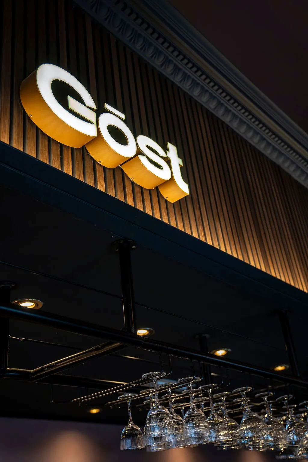 This detail shot captures the sophisticated branding and overhead storage of a hospitality space. A prominent, backlit 3D "Göost" sign with a warm amber glow stands out against a rich, dark fluted wood panel wall, topped by an ornate classic molding. Below, a dark ceiling or soffit features subtle recessed downlights, illuminating an industrial-style black metal rack neatly holding rows of sparkling wine glasses, hinting at a well-appointed bar area. The overall mood is warm, inviting, and subtly dramatic, perfect for an evening setting.