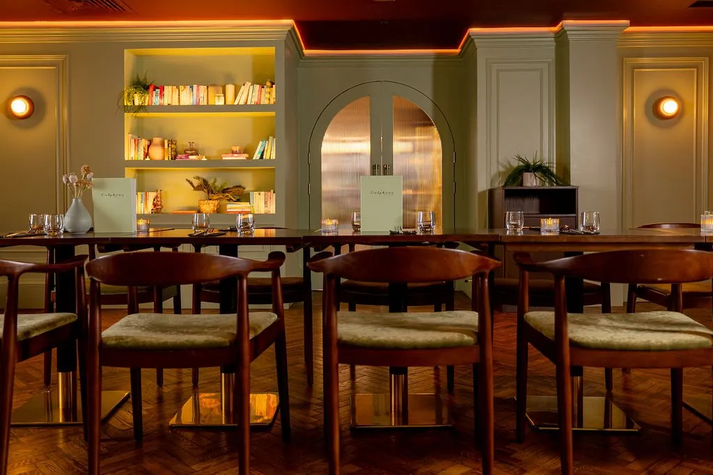 A warm and inviting dining room features sage green paneled walls, a built-in bookshelf, and dark wood tables set with elegant place settings under ambient lighting.