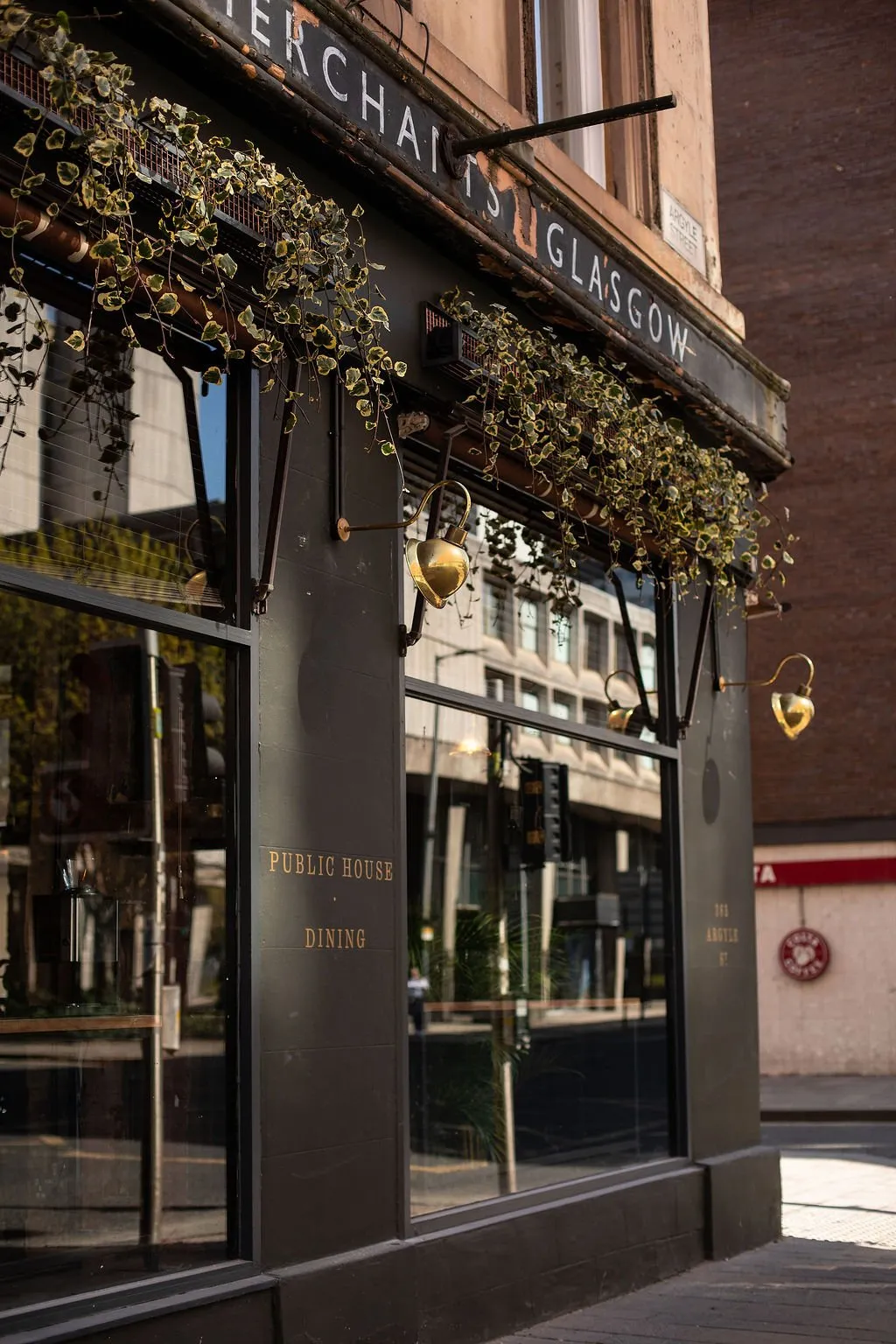 The image captures the sophisticated exterior facade of "Merchants Glasgow," a public house and dining establishment. The dark, possibly charcoal, painted storefront features large reflective windows and elegant gold/brass lettering for its name and services. Above the windows, planters overflow with lush, variegated ivy, complemented by decorative brass wall sconces that add a touch of classic warmth. Bright daylight casts sharp shadows, highlighting the architectural details and creating a welcoming yet refined street presence.