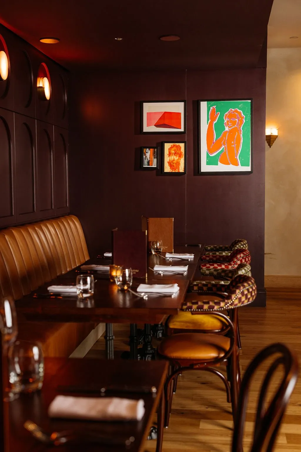 This intimate dining space features a long, channel-tufted tan leather banquette set against a rich plum-colored wall with dark wood arched paneling. Warm, ambient lighting from recessed ceiling lights and decorative wall sconces creates a sophisticated and cozy evening atmosphere. Dark wooden tables are meticulously set with simple place settings and votive candles, complemented by a mix of patterned upholstered chairs and classic bentwood-style seating, all beneath an eclectic gallery wall of vibrant modern art.